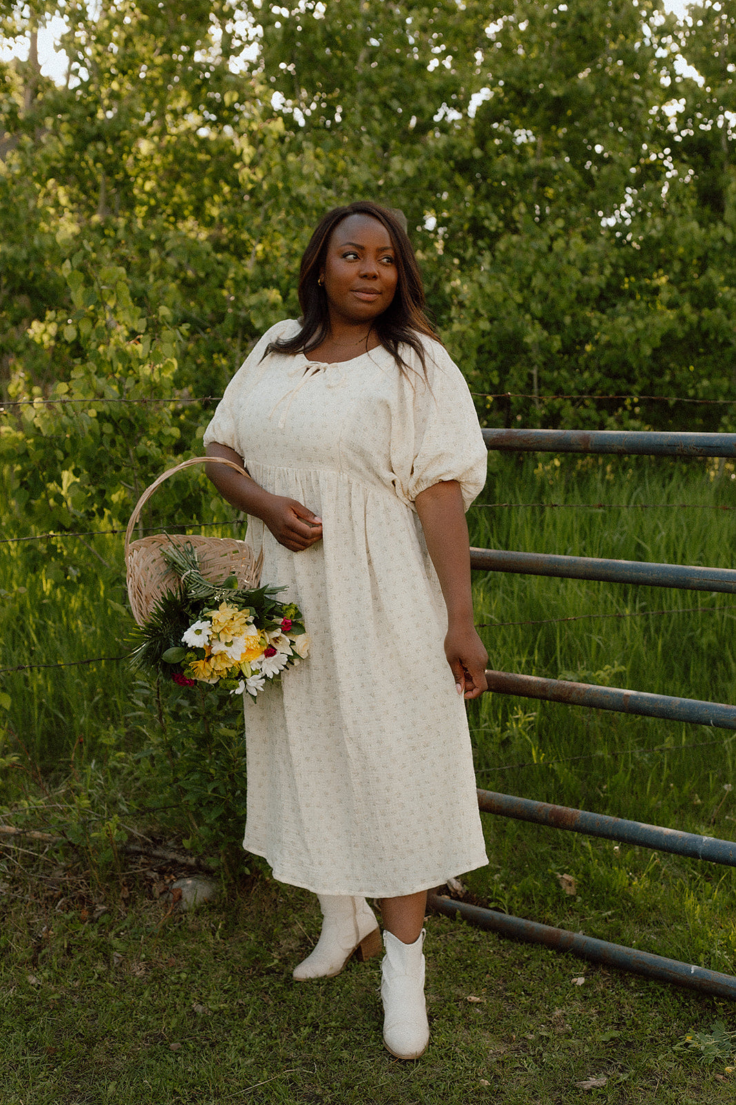 A woman stands outdoors in a grassy field wearing a soft floral yellow, short-sleeved midi dress with a subtle floral pattern, holding a wicker basket filled with yellow and white flowers. She is wearing white ankle boots and standing in front of a green tree line and a metal fence. its a breastfeeding dress 
