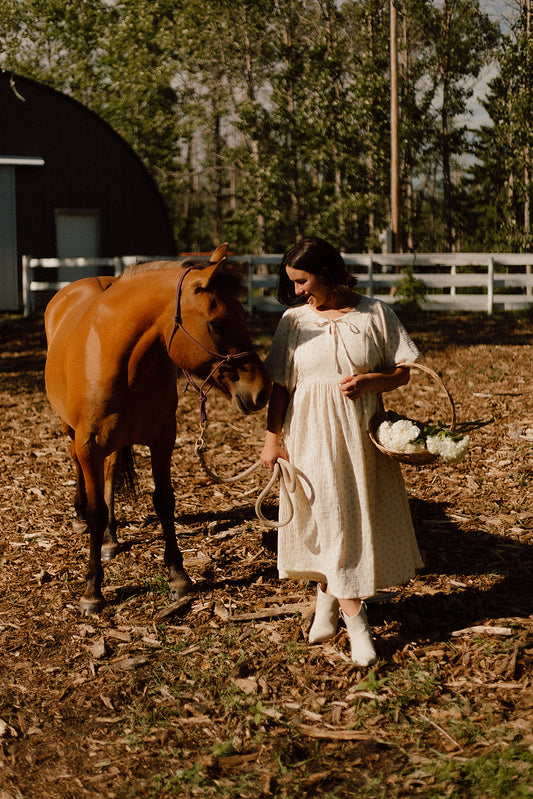 A woman in a cream-colored vintage-style dress stands in an outdoor barnyard next to a brown horse, holding a woven basket of white flowers.