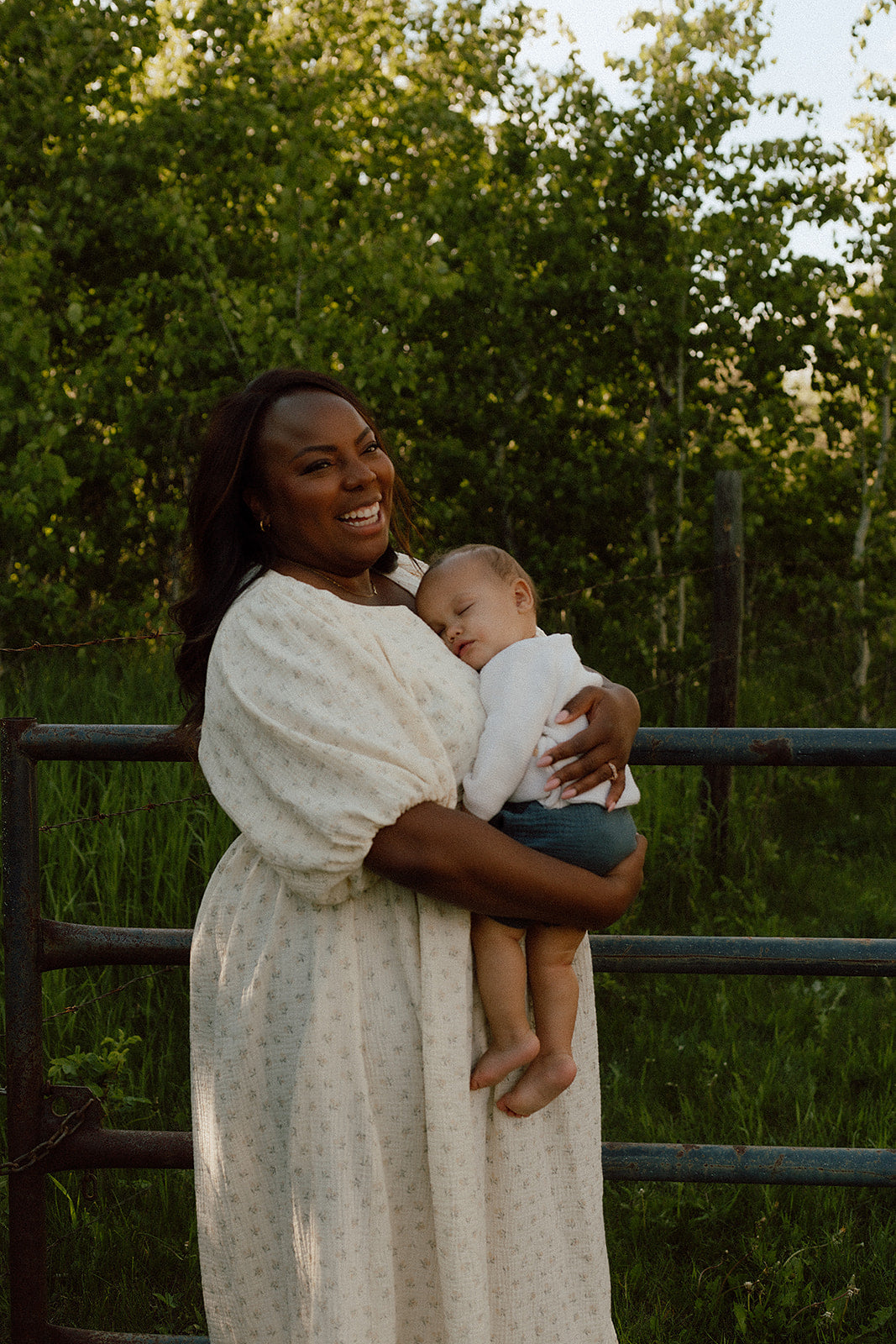A smiling woman outdoors holding a sleeping baby, wearing a cream-colored long-sleeve dress with a subtle small dot pattern. The background features lush green trees and a dark metal fence.