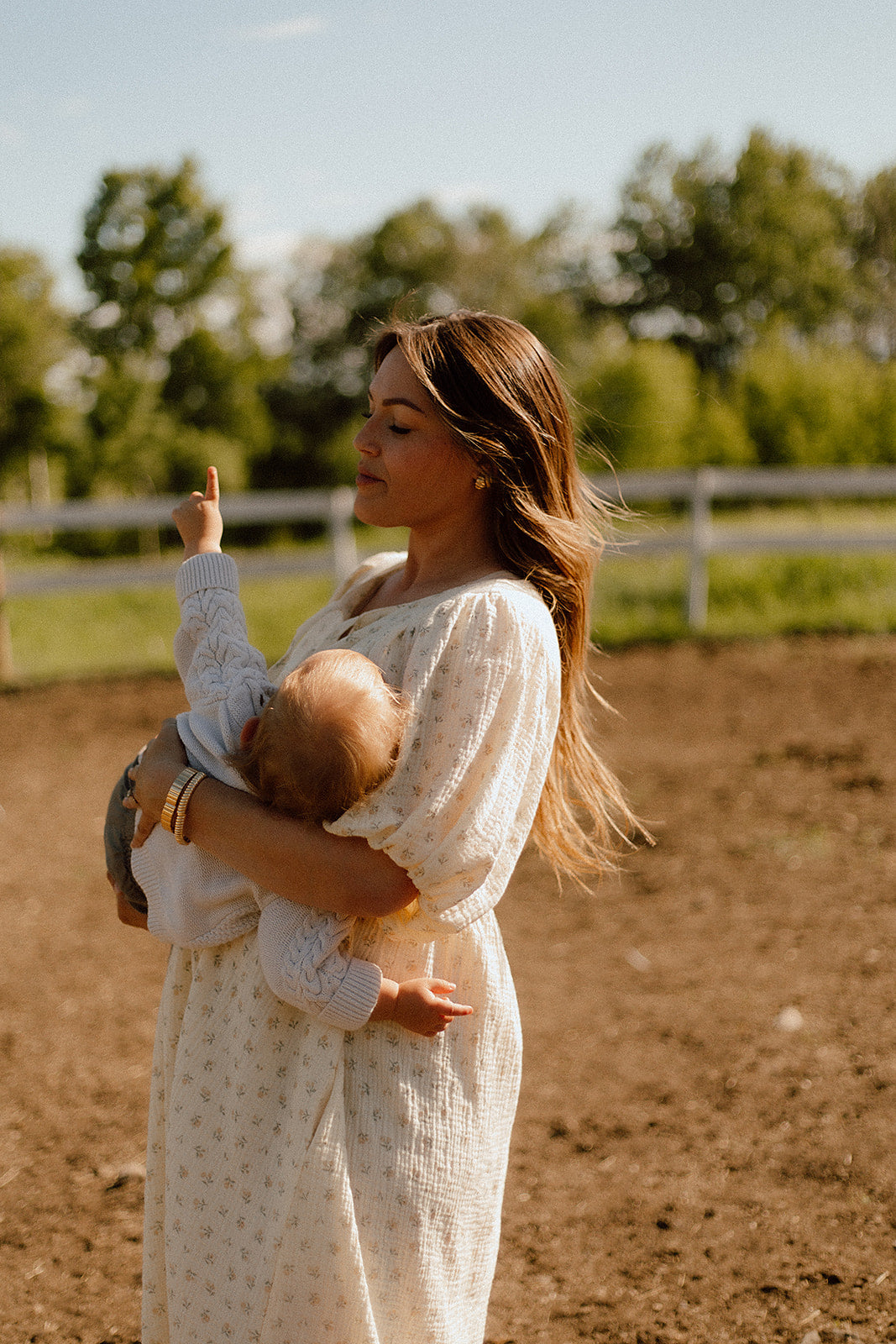 A woman in a white long-sleeve babydoll dress holds a baby in an outdoor dirt paddock with trees and a fence in the background.