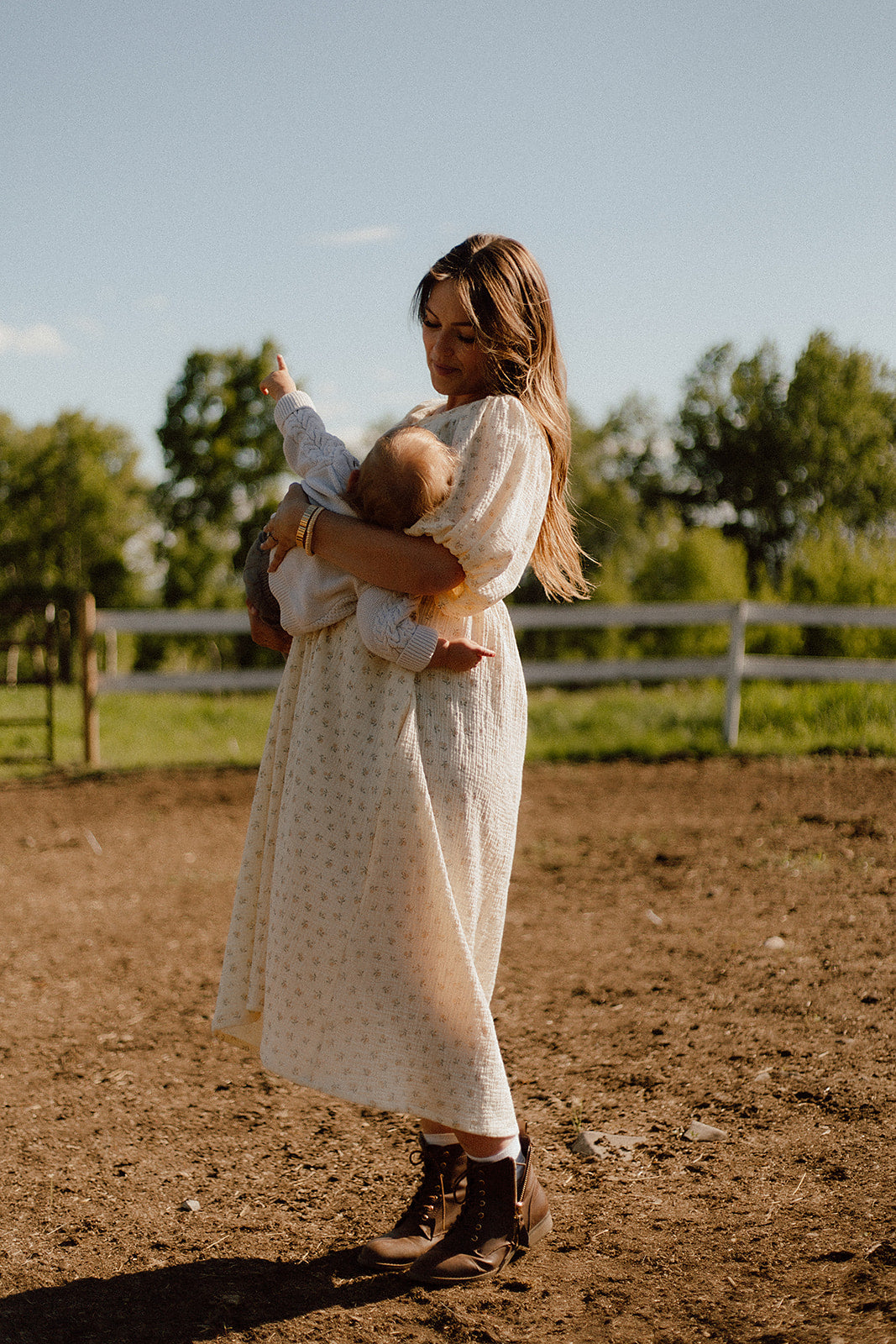 A woman in a cream breastfeeding babydoll dress with a floral pattern holds a baby outdoors in a dirt paddock with a white fence and green trees in the background.