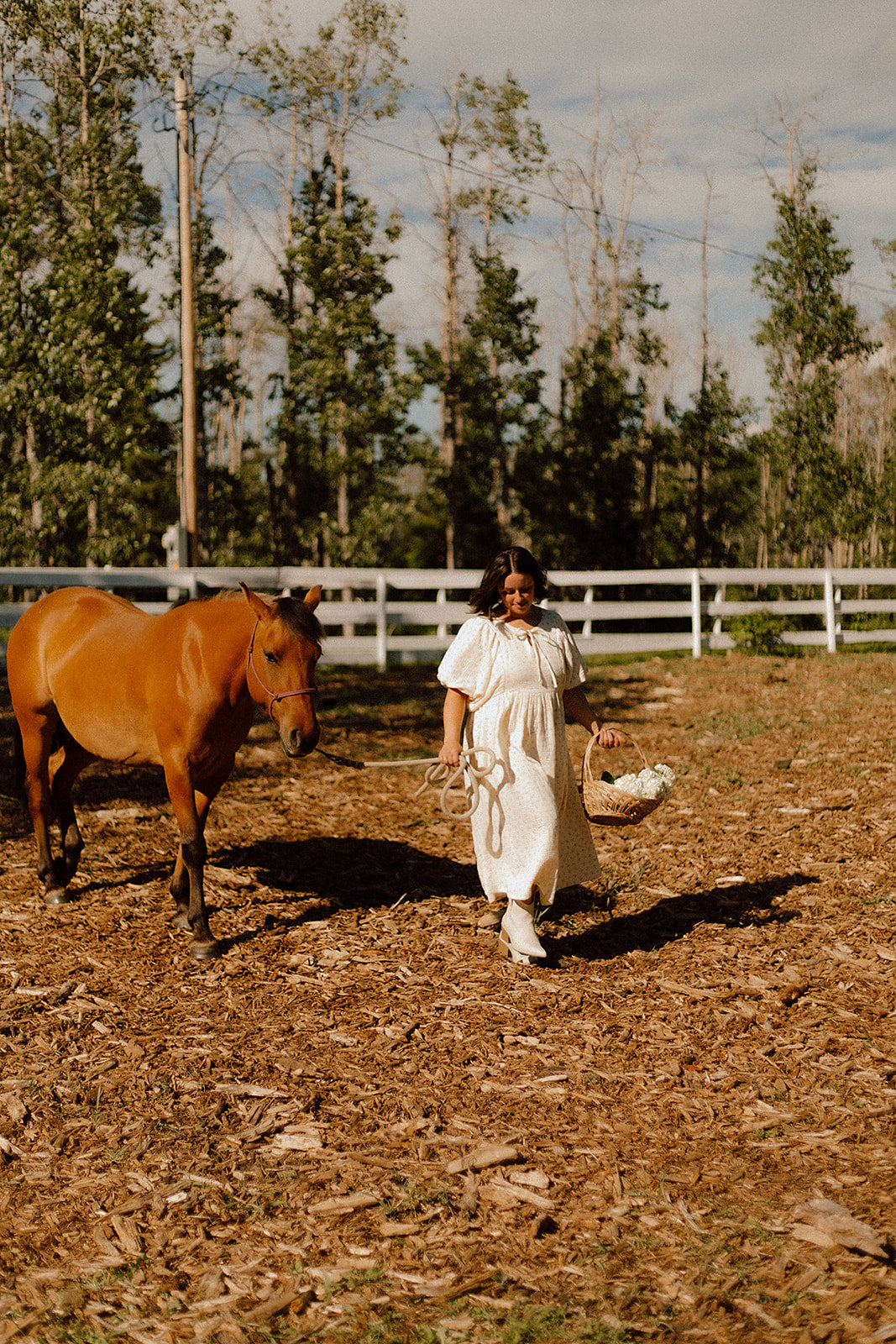 A woman in a yellow floral breastfeeding dress walks in a dirt paddock with a brown horse beside her, with a white fence and tall trees in the background.